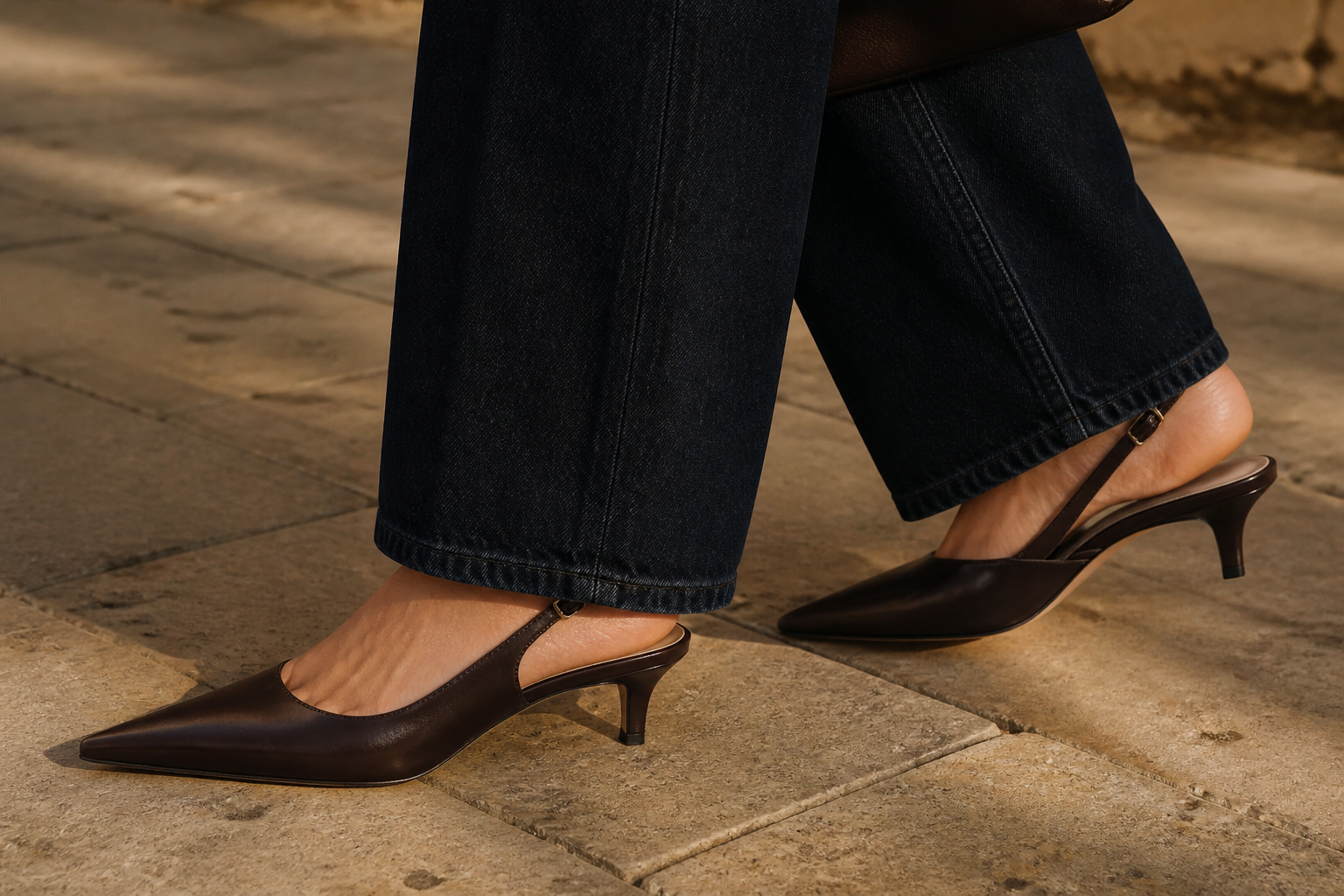 Close detail of espresso kitten-heel slingbacks under dark straight-leg denim on warm stone pavement
